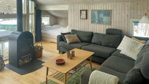 A cozy cabin interior with a wooden ceiling, a built-in bed, a gray sofa, a small table, and a fireplace. - Cottage with beach access on Sjællands Odde (Sjællands Odde)