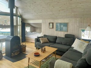Ein gemütliches Blockhauskaminzimmer mit Holzbalkendecke, integriertem Bett, grauem Sofa, kleinem Tisch und Kamin.