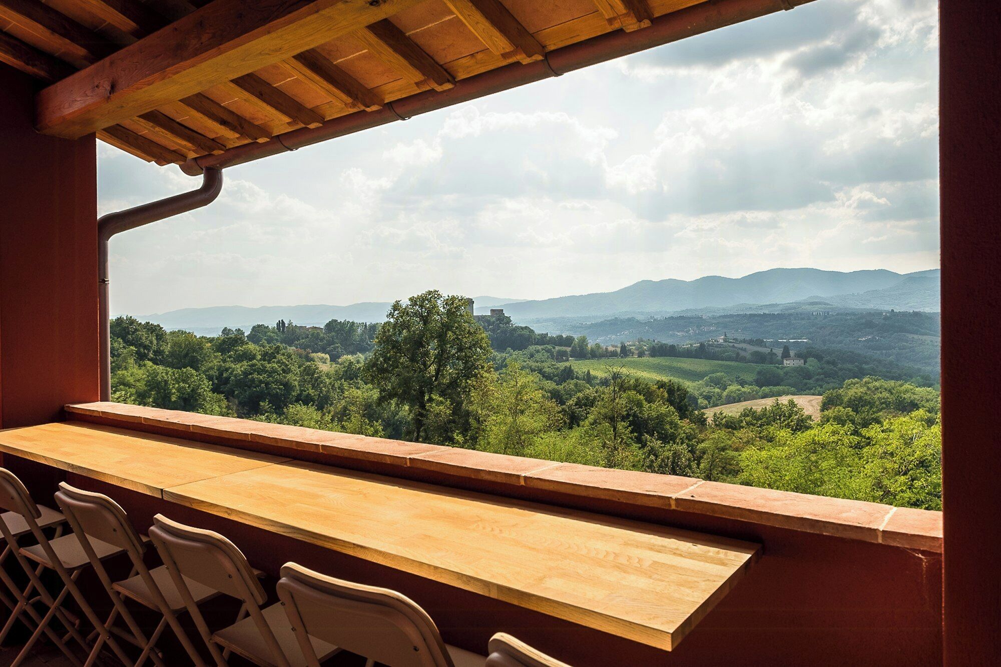 A wooden bar with chairs, a view of a green landscape with trees and hills, and a cloudy sky.