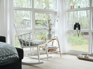 Ein Zimmer mit Schaukelstuhl, einem kleinen Tisch mit Blumenvase und einem Fenster mit Fernglas auf der Fensterbank.