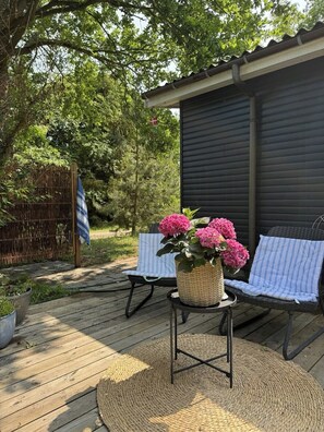 Eine Holzterrasse mit einem Korbstuhl-Tisch, auf dem ein Blumenkorb steht, zwei gestreifte Lounge-Stühle und ein Gebäude im Hintergrund.