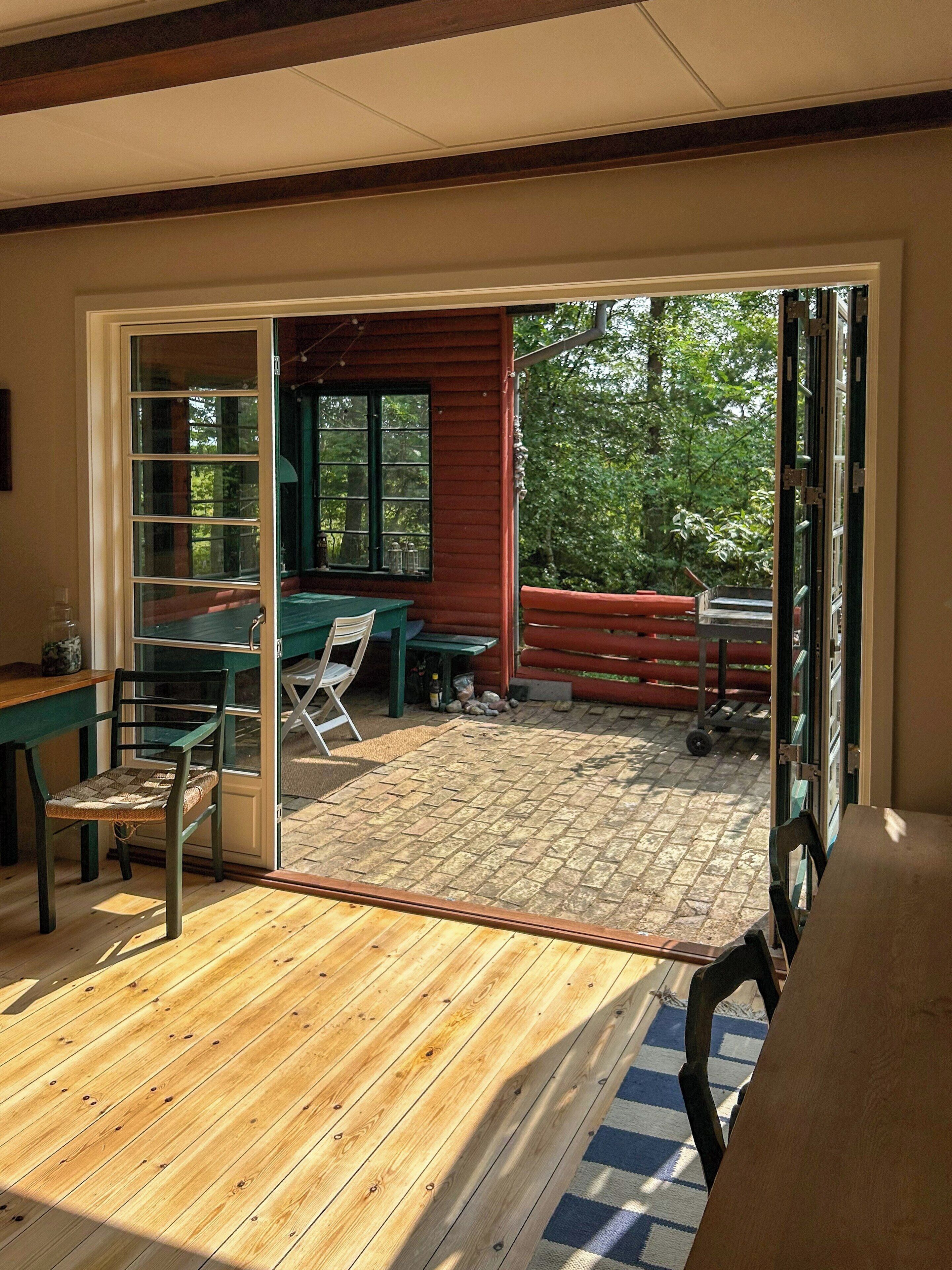 Een houten terras met een tafel en stoelen, een schuifdeur naar een buitenruimte met een bankje en een raam.