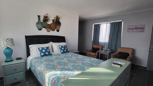 Une chambre d’hôtel comprenant un lit, une table de chevet, une commode, une chaise, une petite table et une fenêtre avec des rideaux.