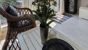 Una habitación con una silla de mimbre, una mesa blanca, un tazón negro, una planta en maceta, un mueble negro y un televisor.