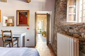 A narrow hallway with a bed, a desk, a chair, a radiator, and a window. - Medieval stone house in the Garfagnana hills (San Romano in Garfagnana)