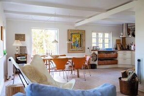 Ein geräumiger Wohnbereich mit Esstisch, orangefarbenen Stühlen, einem weißen Sofa und einer Küchenzeile mit Herd und hängenden Töpfen.