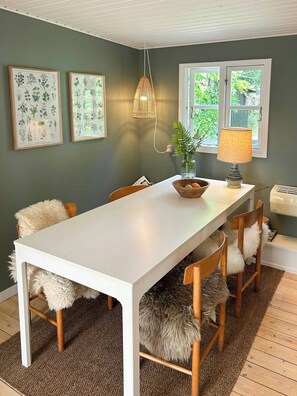 A dining area with a white table, wooden chairs, a bowl, a lamp, and framed botanical illustrations on the wall. - Forest house with private grounds in Liseleje (Frederiksværk)
