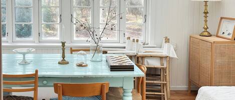 A room with a dining table, chairs, a lamp, a bed, and a window with a view of trees.