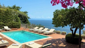 Zona de piscina con sillones de descanso, vista despejada al mar y un árbol con flores rosas.