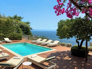 Ein Poolbereich mit Liegestühlen, freier Meeresblick und ein Baum mit rosa Blüten.