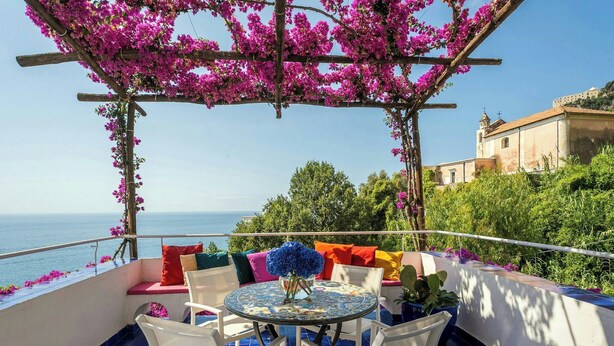 Una terraza con mesa y sillas, una pérgola adornada con flores y vistas al mar y a un edificio.