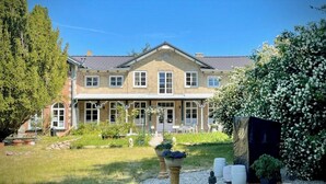 Ein zweistöckiges Haus mit einer symmetrischen Fassade, einer Veranda und einem gepflegten Garten mit einer Statue, Topfpflanzen und einem Strauch.