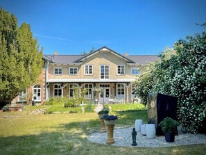 Ein zweistöckiges Haus mit einer symmetrischen Fassade, einer Veranda und einem gepflegten Garten mit einer Statue, Topfpflanzen und einem Strauch.