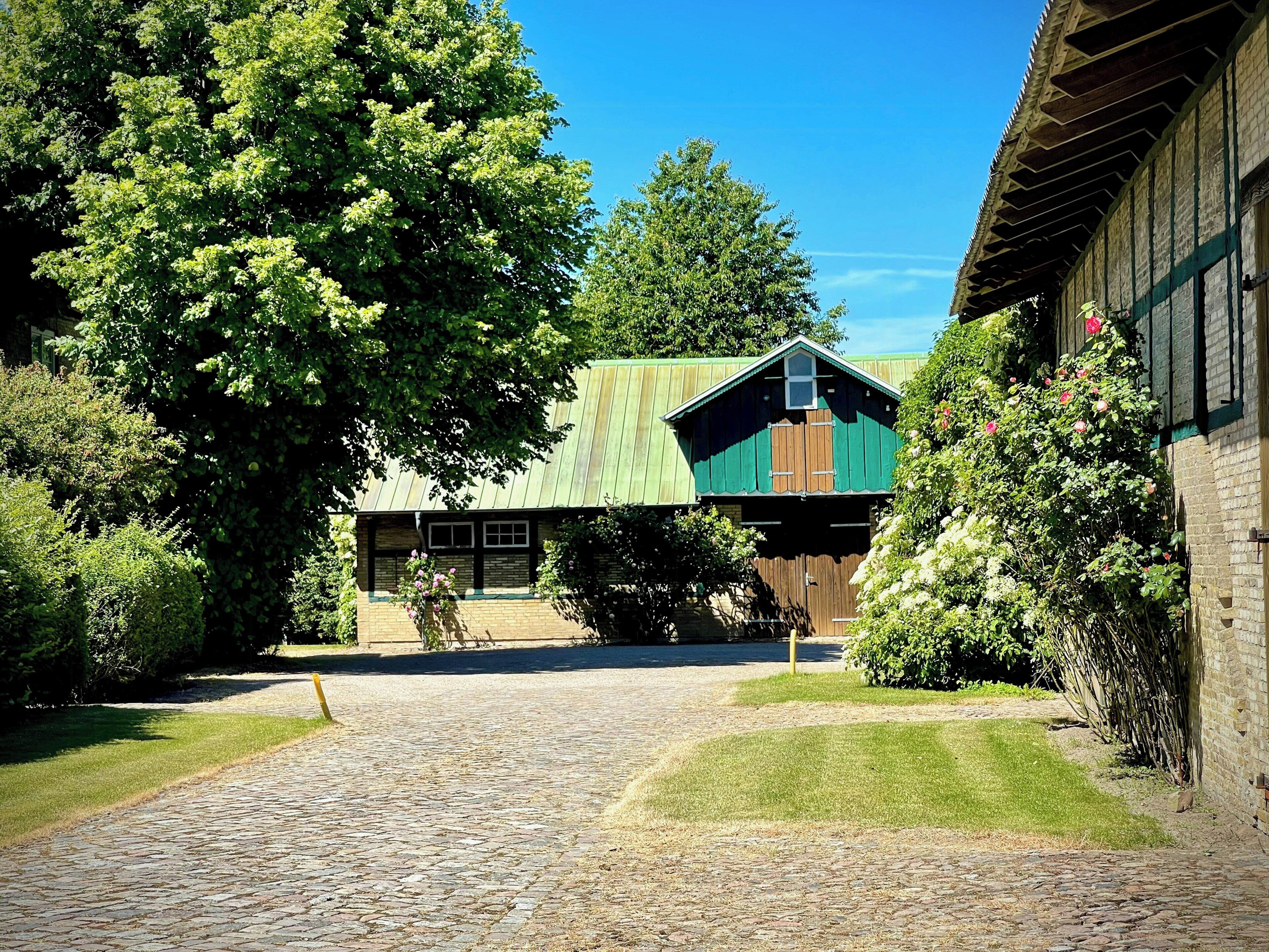 A cobblestone pathway leading to a building with a green roof and wooden walls, surrounded by lush greenery.