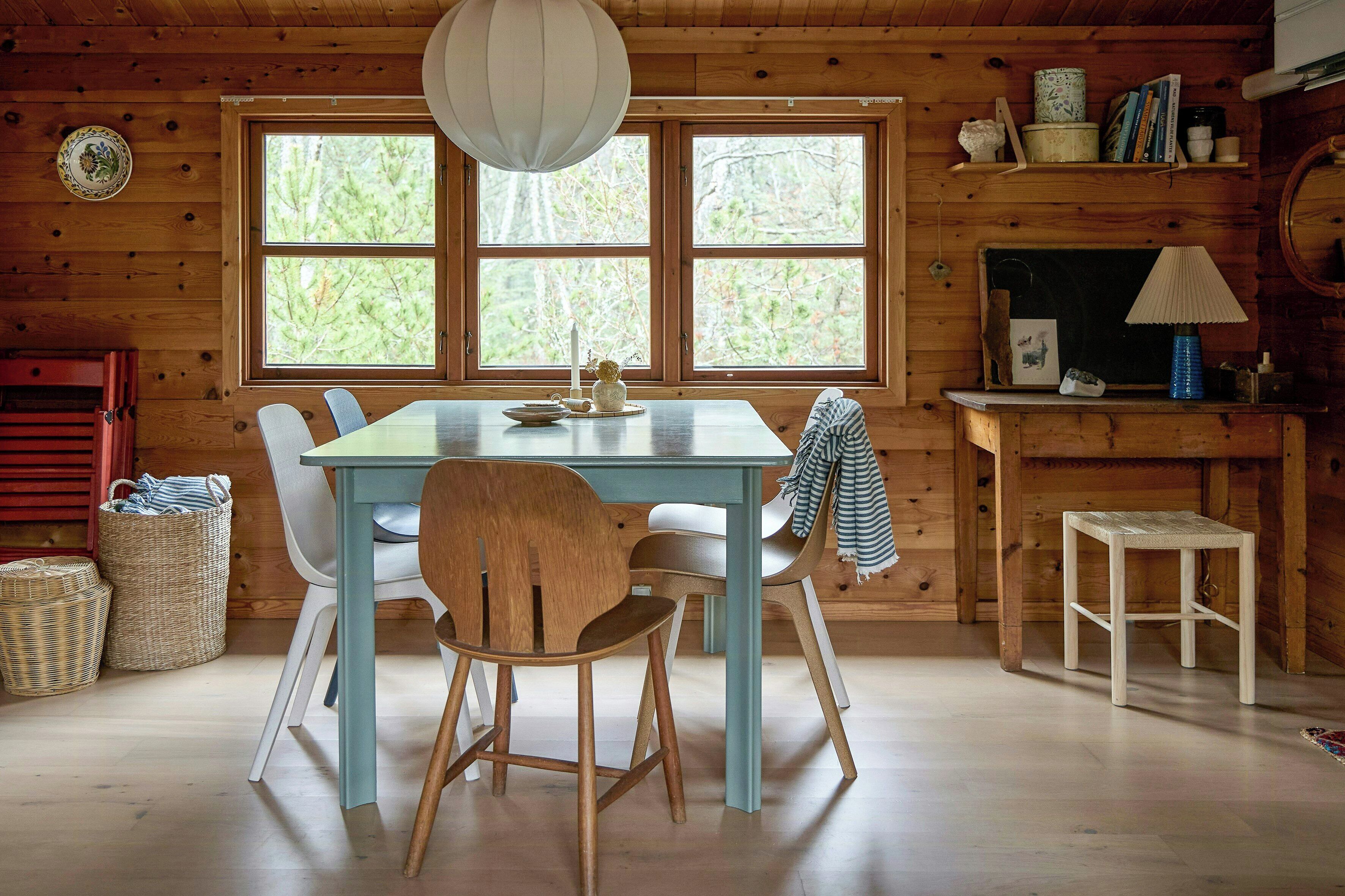 Interior de madeira com uma mesa de jantar, cadeiras, luminária pendente e janela com vista para árvores.