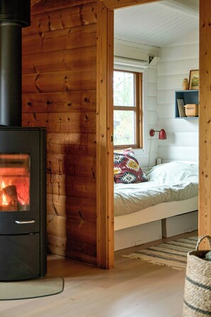 A cozy cabin interior with a wood-burning stove, a bed with a colorful pillow, and a window with a view of trees. - Cozy gem near Tversted Plantage (Aalbæk)