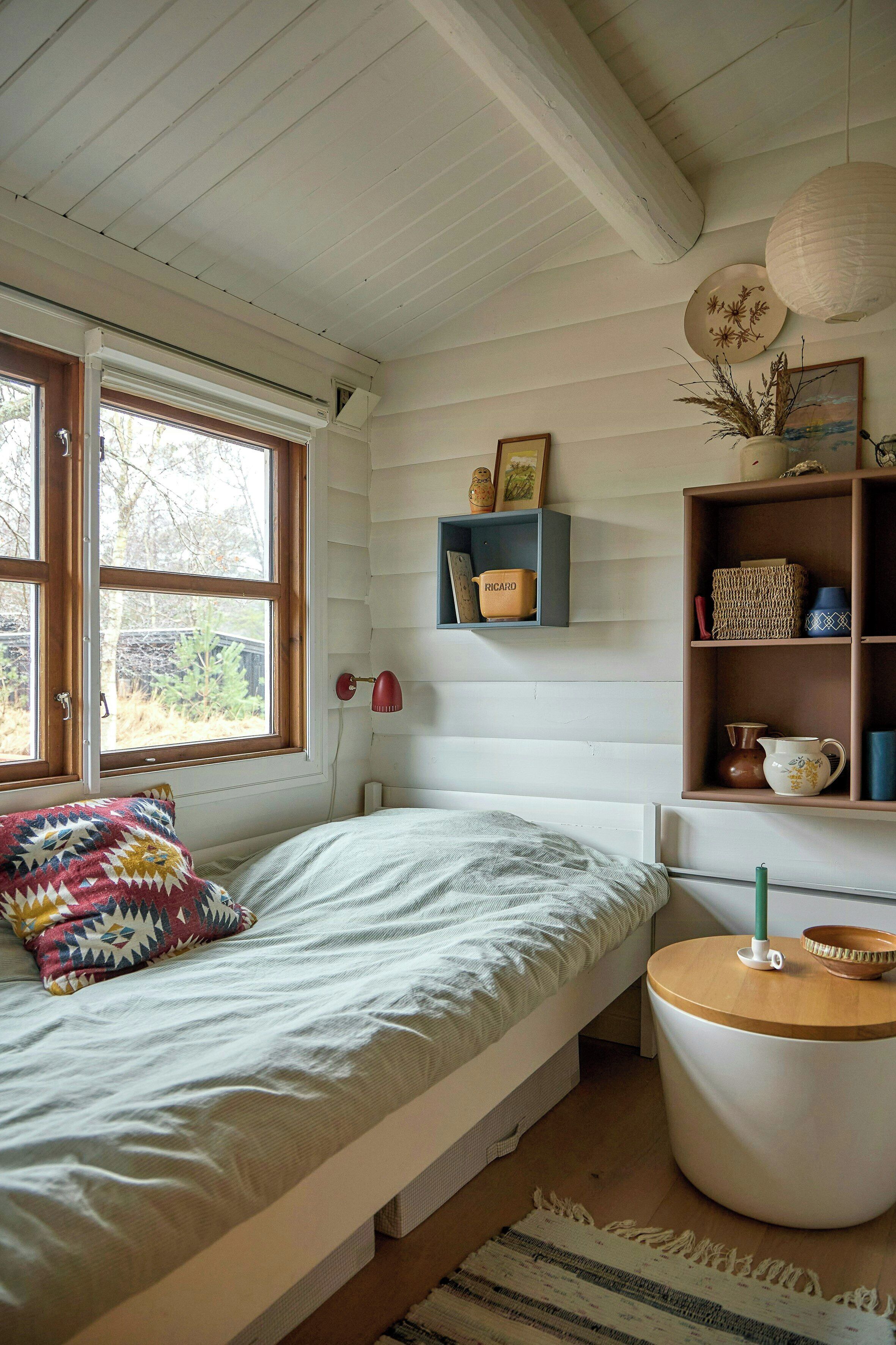 Un dormitorio con cama, mesita de noche, lámpara, estante con adornos y ventana.