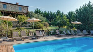 Ein Schwimmbecken mit Liegestühlen und Sonnenschirmen, umgeben von einer Holzterrasse und einem Steinhaus.