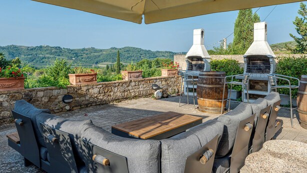 Eine Terrasse im Freien mit einer Steinmauer, einem Holztisch, einem Grill und Blick auf die Hügel.