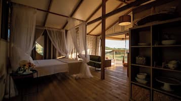 A room with a bed, a small table with a fruit bowl, a shelf with dishes, and a view of the outdoors.