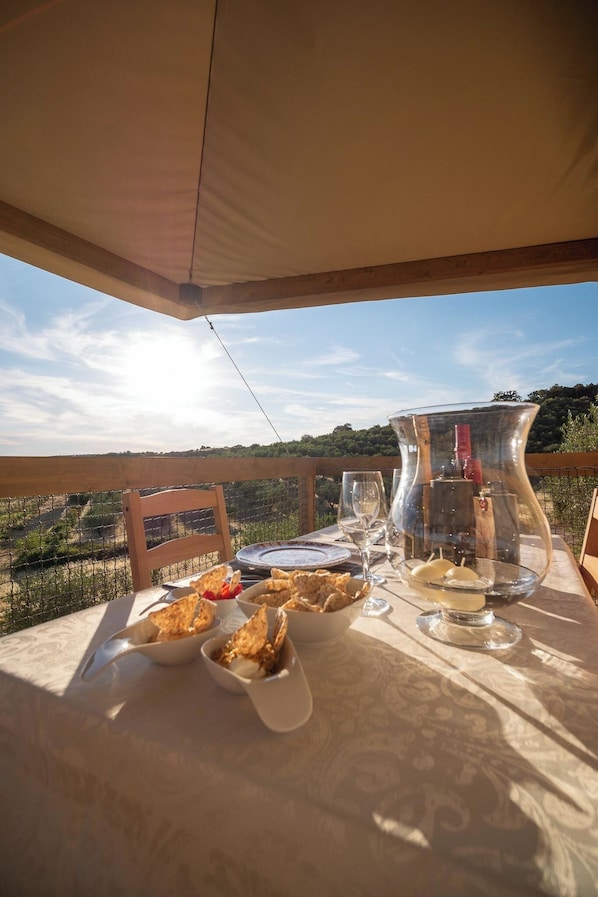 An outdoor dining table with food and drinks, a wooden chair, and a view of the landscape.
