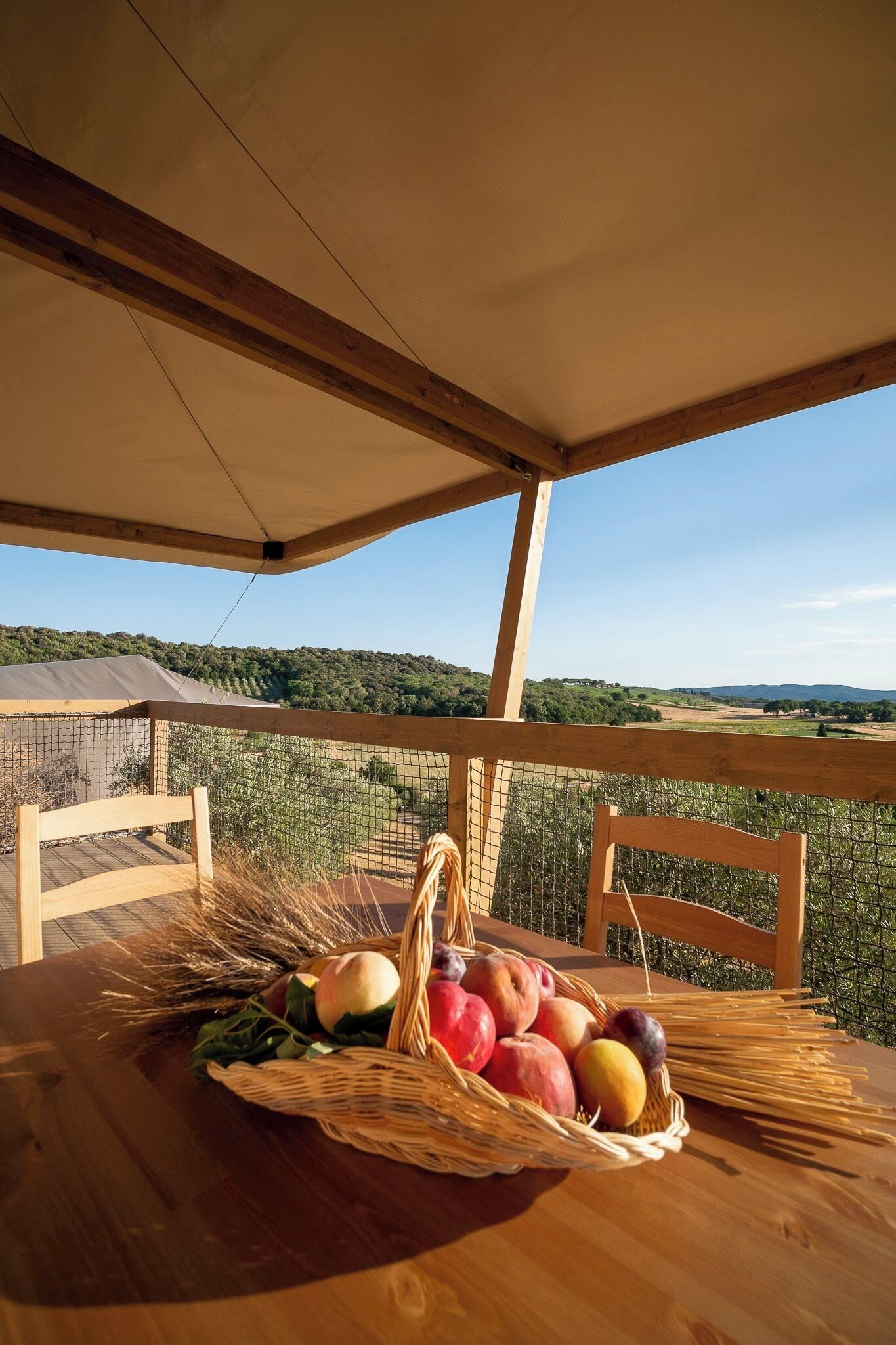 Una mesa de madera con una cesta de frutas y verduras, una silla de mimbre y vistas a un paisaje montañoso.