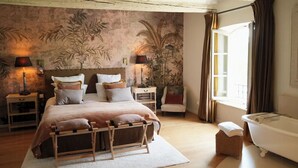 Un dormitorio con cama, mesitas de noche, lámpara, sillón, ventana y bañera.