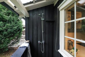 Un mur extérieur noir avec une fenêtre, une pomme de douche en métal et une petite table avec des chaises à l’extérieur.