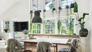 A spacious room with a large wooden table, multiple chairs, a television, and a variety of plants. - Step through the gate and feel peace in the woods (Silkeborg)