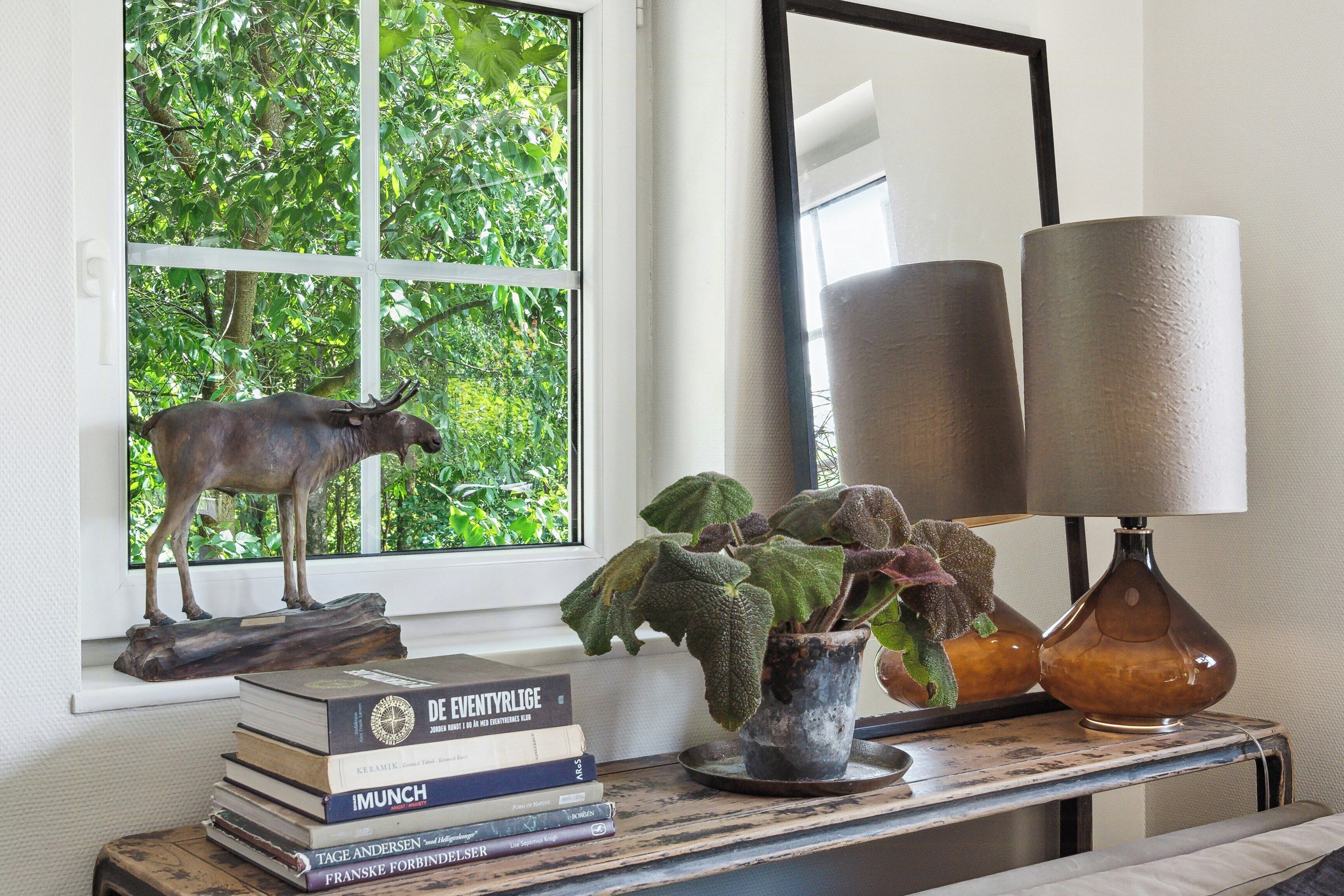 A window with a view of greenery, a statue of a bull, a stack of books, a potted plant, and a lamp on a table.