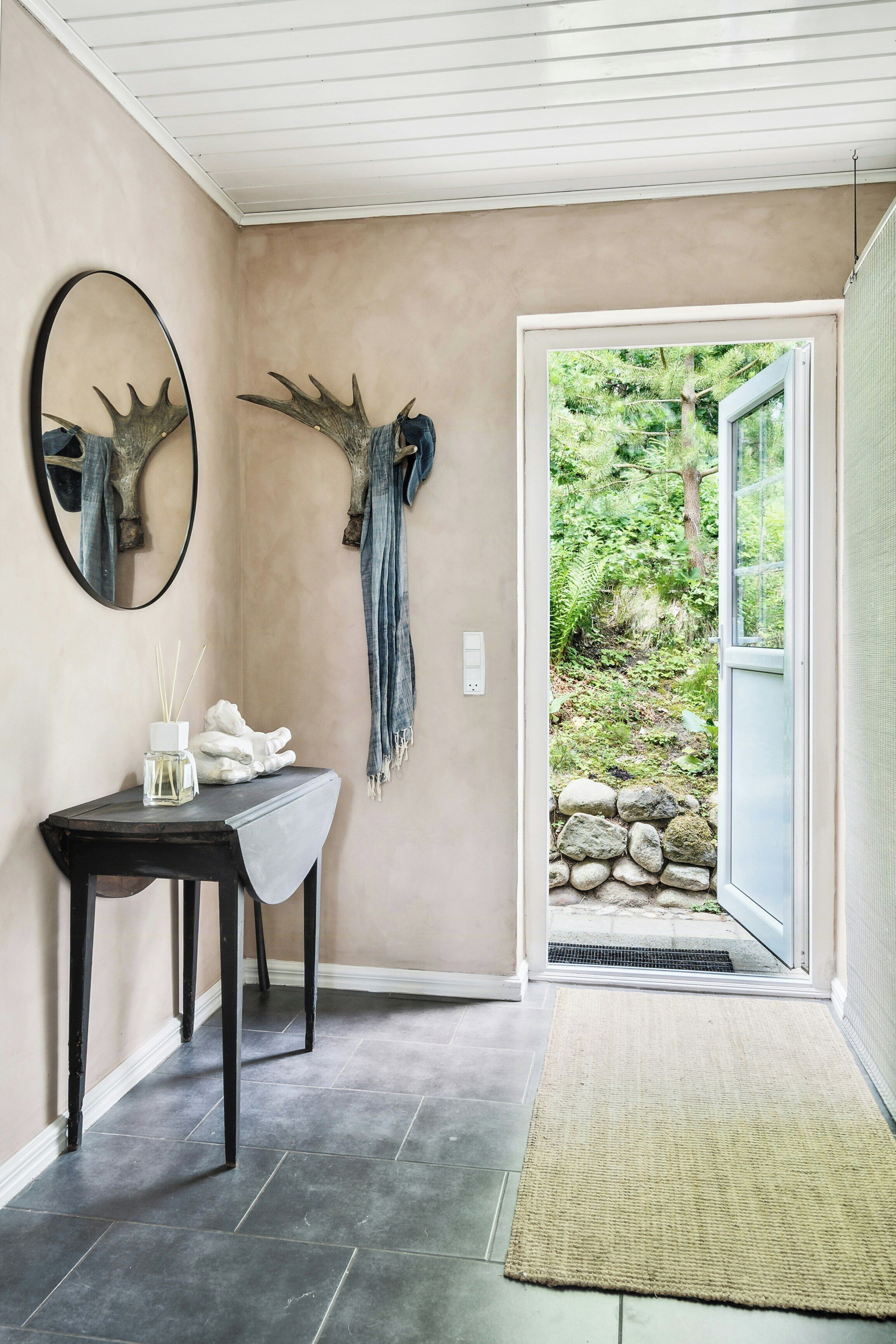 A room with a stone wall, a mirror with antlers, a small table with a bottle, and a view of greenery outside.