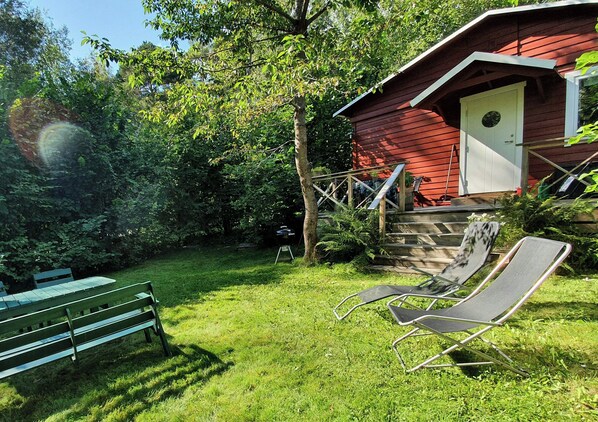 A red wooden house with a white door and steps leading up to it. There is a green bench and a black lounge chair in the yard. - Cottage by the sea on Djurö (Djurhamn)