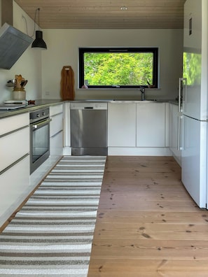 Eine moderne Küche mit weißen Schränken, einem Edelstahl-Geschirrspüler, Holzboden und einem Fenster mit Blick auf Grünflächen.