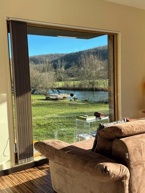 Ein Zimmer mit einer großen Schiebeglasdoor zum See, einem Sofa und einem kleinen Tisch mit Büchern.