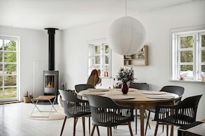 Een eetgedeelte met een ronde tafel, zwarte stoelen, een witte hanglamp en een houtkachel.
