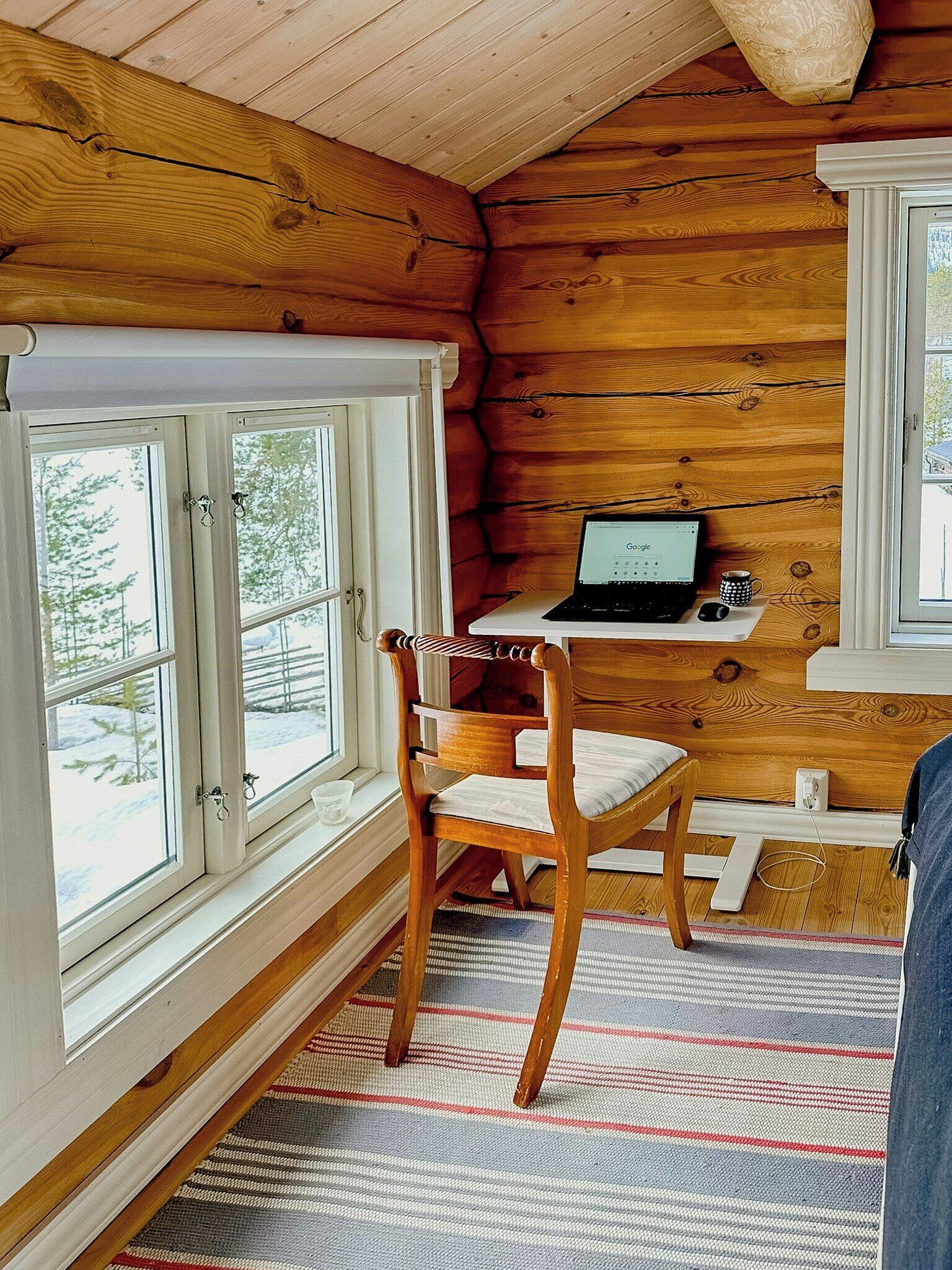 A cabin interior with a wooden desk, a laptop, a chair, and a window with a view of trees.