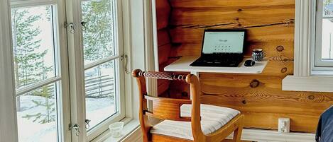 A cabin interior with a wooden desk, a laptop, a chair, and a window with a view of trees.