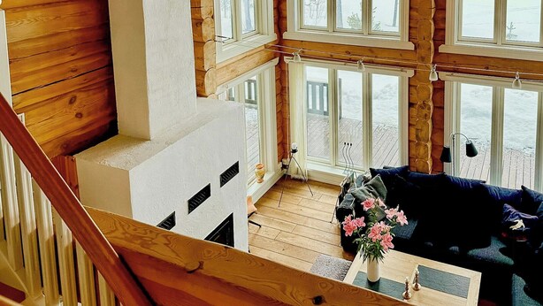 A cabin interior with a wooden staircase, a fireplace, a wooden deck visible through large windows, and a dark-colored sofa.