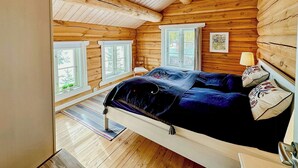 A log cabin bedroom with a bed, windows, a lamp, and a framed picture on the wall.