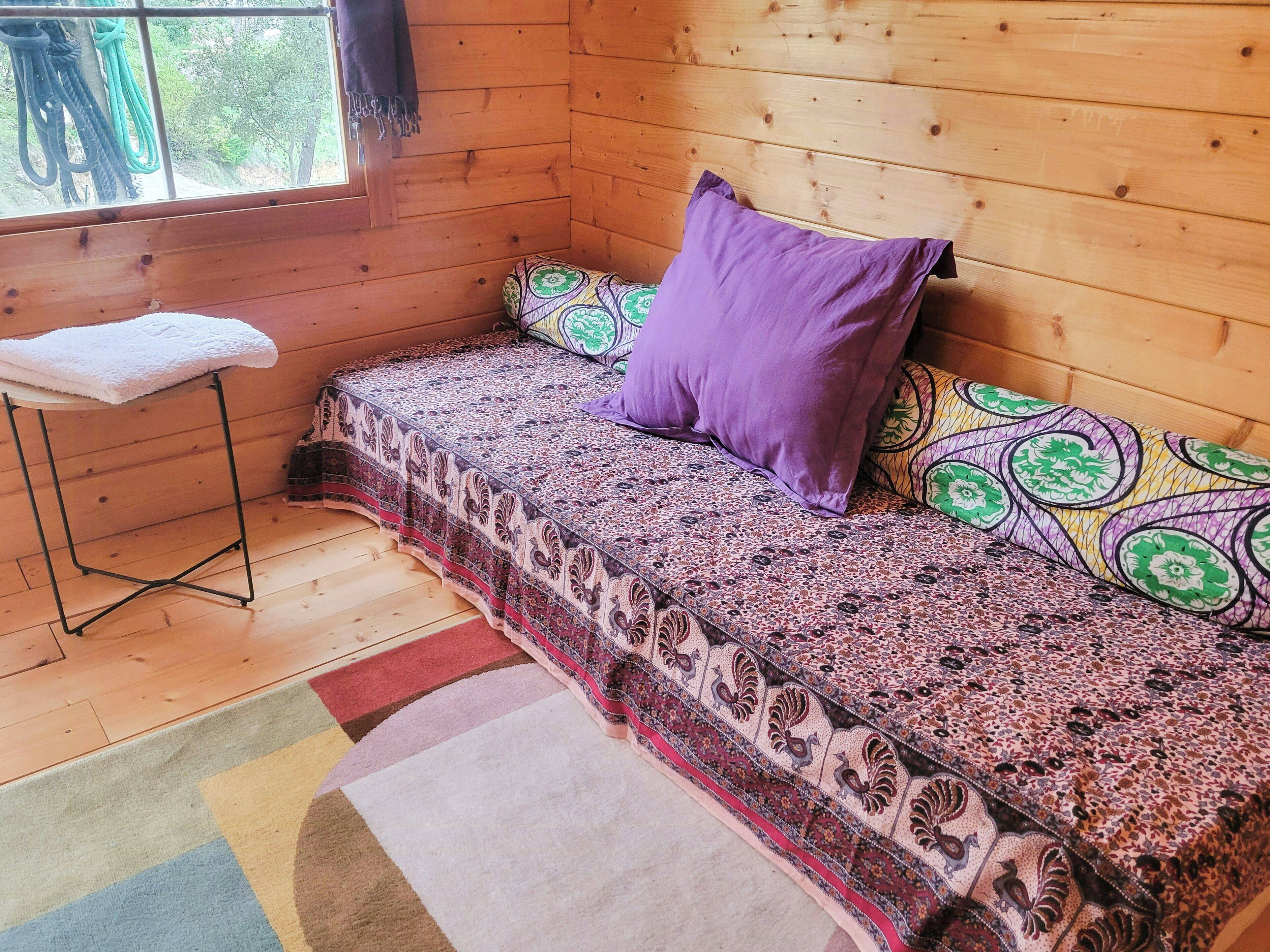 Una habitación de cabaña acogedora con interior de madera, una cama con ropa de cama estampada, un taburete pequeño con una toalla y una alfombra colorida.