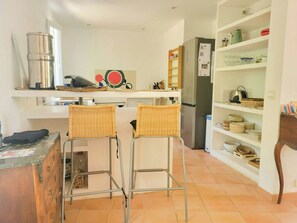 Eine Küche mit zwei Barhocker, einem Kühlschrank, Regalen mit Geschirr und einem Holztisch.