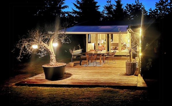 Eine Holzterrasse mit Esstisch und Stühlen, umgeben von Kübelpflanzen und einem großen Baum.