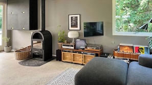 Ein Wohnzimmer mit Kamin, Fernseher, Sofa und einem Fenster mit Blick ins Grüne.
