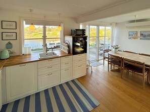 A modern kitchen with a large window, a dining area with a table and chairs, and a view of a deck and trees. - Holiday home by the water in Øer Maritime (Ebeltoft)