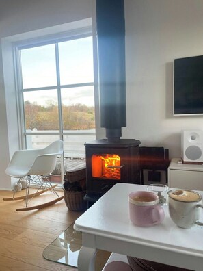 A room with a wood-burning stove, a white rocking chair, a table with a glass top, and a white wall-mounted speaker. - Holiday home by the water in Øer Maritime (Ebeltoft)