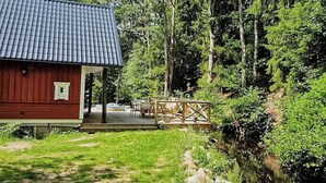 Una baita di legno rossa con tetto in tegole, immersa in un prato verde e attraversata da un piccolo ruscello.