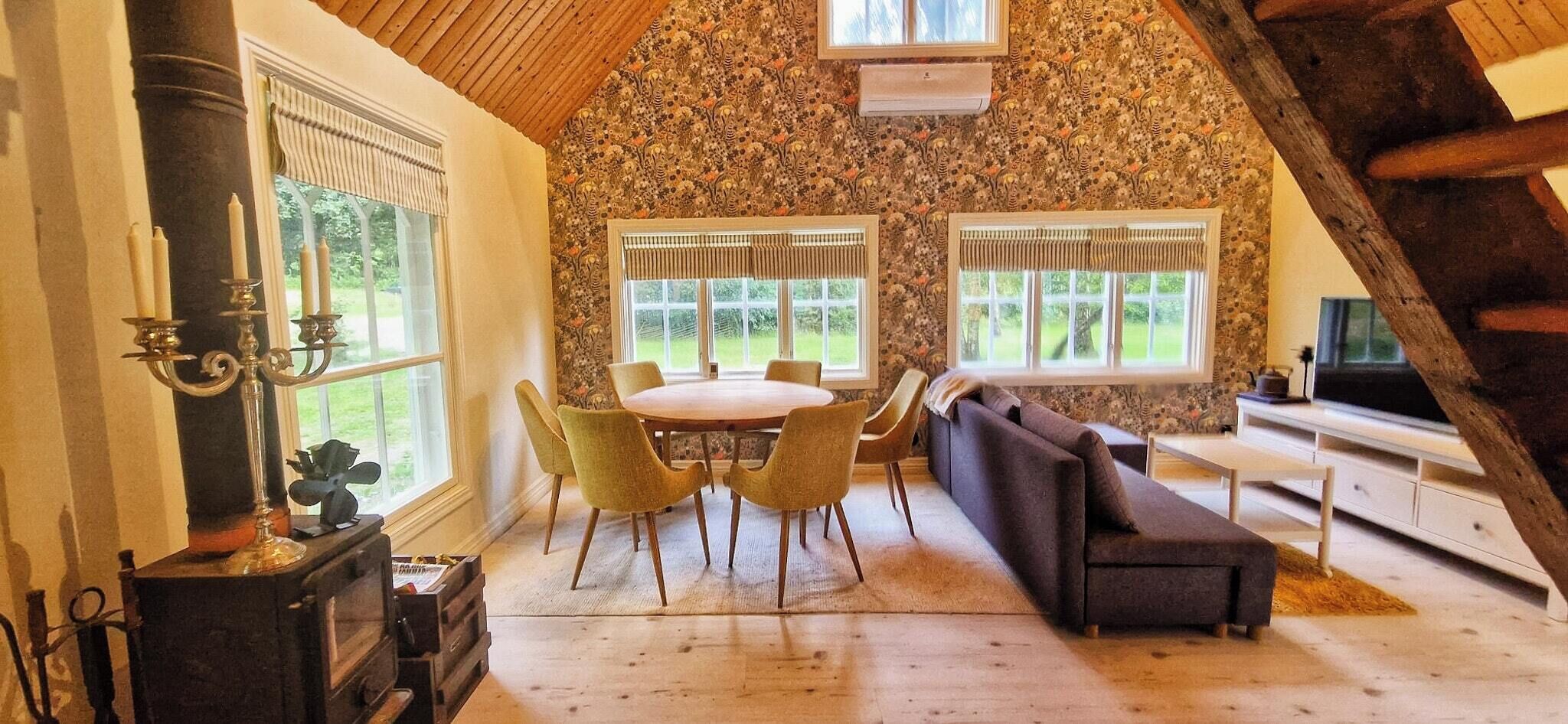 Une pièce chaleureuse avec un plafond en bois, une table ronde entourée de chaises, un canapé et deux fenêtres avec des rideaux.