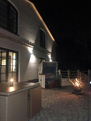 A building with a white exterior, a wicker chair, a small table with a candle, and a fire pit.
