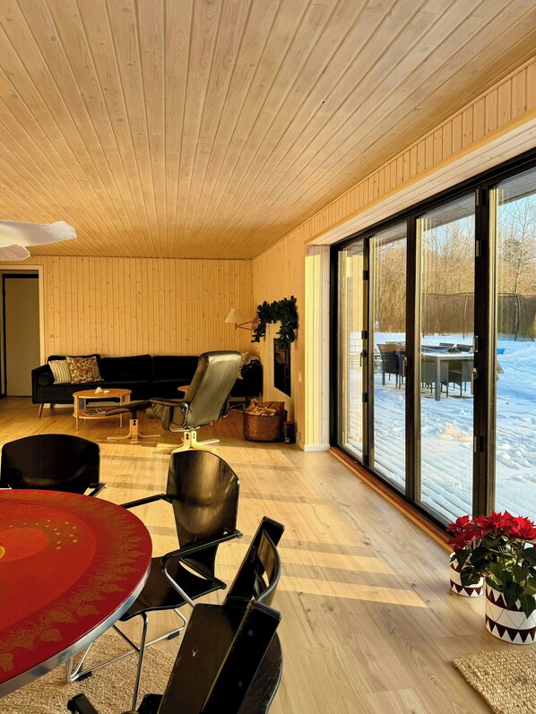 Ein modernes Interieur mit Holzbalkendecke, einem Essbereich mit rotem Tisch, einer schwarzen Couch und einem großen Fenster mit Blick auf eine verschneite Landschaft.
