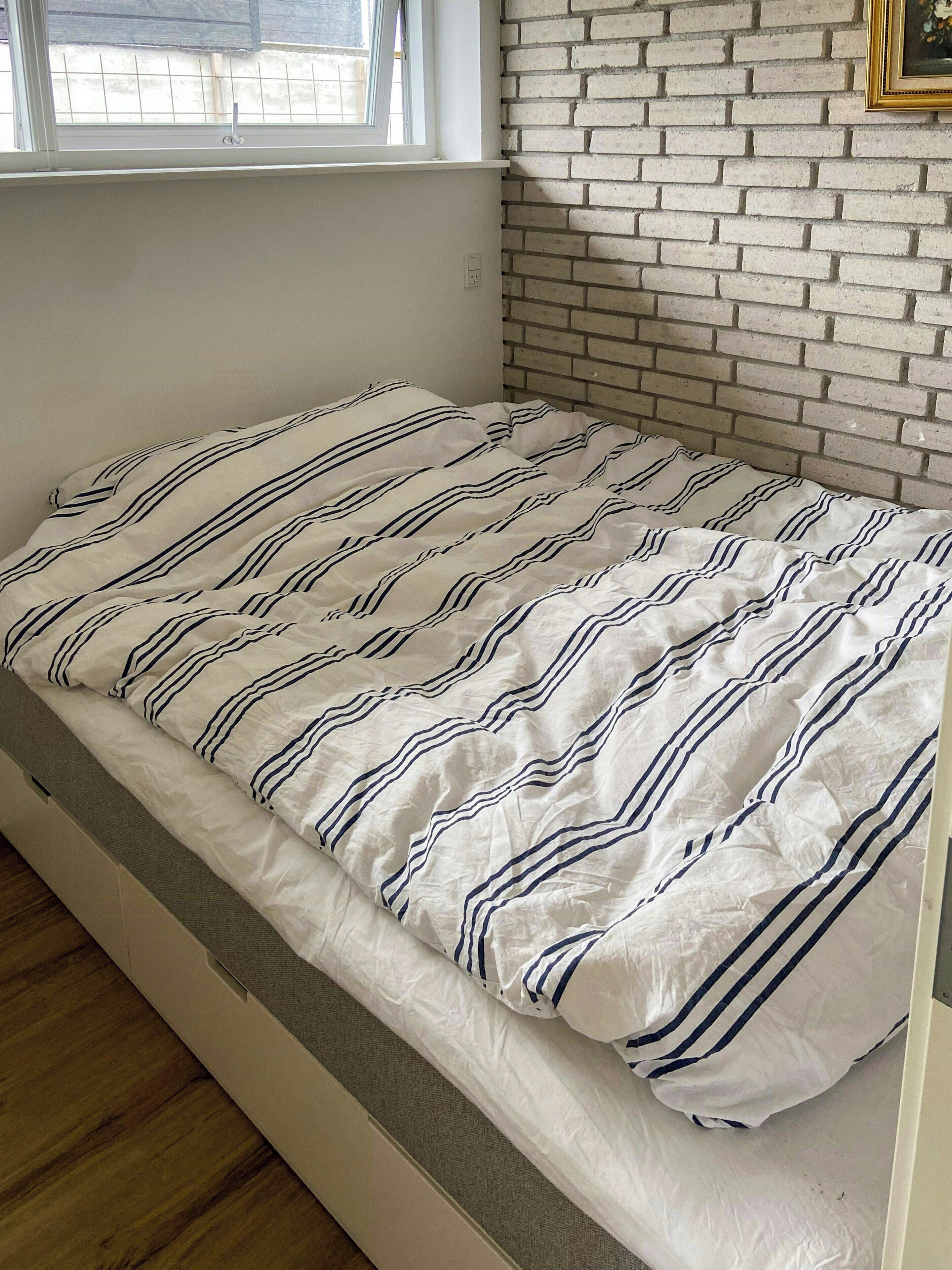 A single bed with a striped bedding pattern, a brick wall, and a window with blinds.